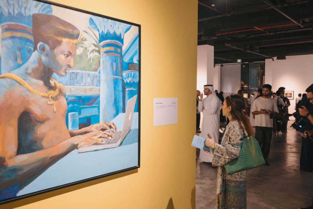 A woman reads a placard next to a painting of an ancient Egyptian figure using a laptop at an art gallery. Other visitors view artworks in the background.