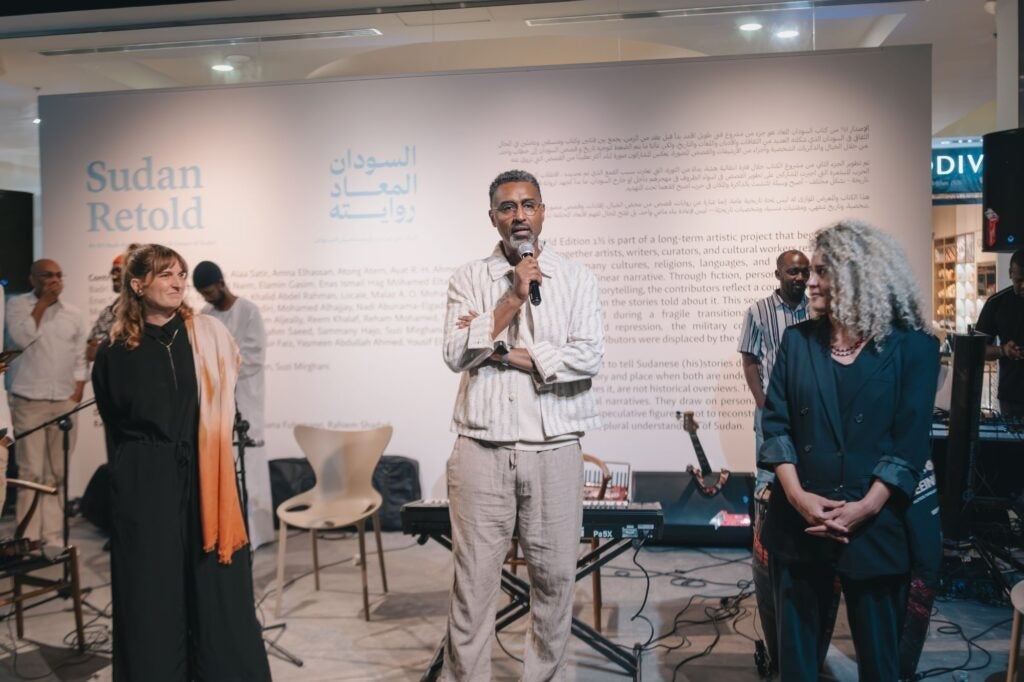 A man speaks into a microphone at an indoor event with the Sudan Retold display behind him. Two women stand nearby, and several people and musical equipment are in the background.
