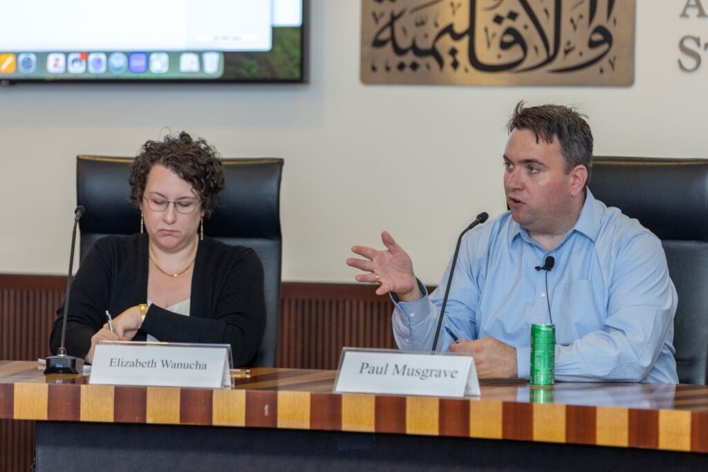 Two people sit at a panel table with microphones. One checks her watch while the other speaks, gesturing with his hand. Name tags read Elizabeth Warnucha and Paul Musgrave. Arabic calligraphy art hangs on the wall behind them.