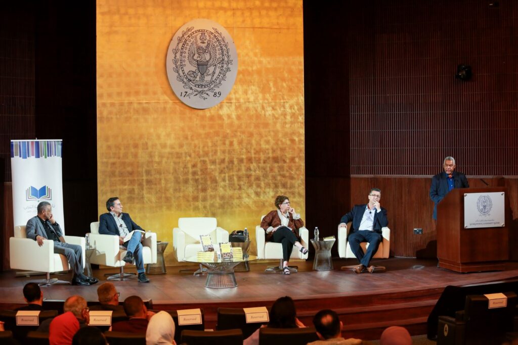 Image of the stage inside the auditorium with 4 panelists and Dr. Abdelwahhab El-Affendi speaking on thepodium