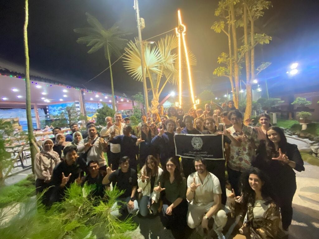 A large group of people pose together outdoors at night, some making hand signs. They stand near tropical plants, and a few hold a black flag with an emblem. The setting is brightly lit and festive.