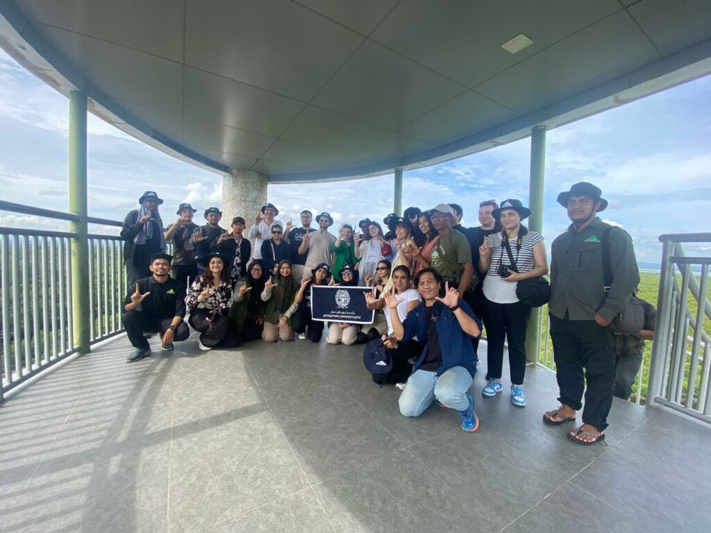 A large group of people poses together on an observation deck under a roof, some kneeling and others standing, with greenery and blue sky in the background.
