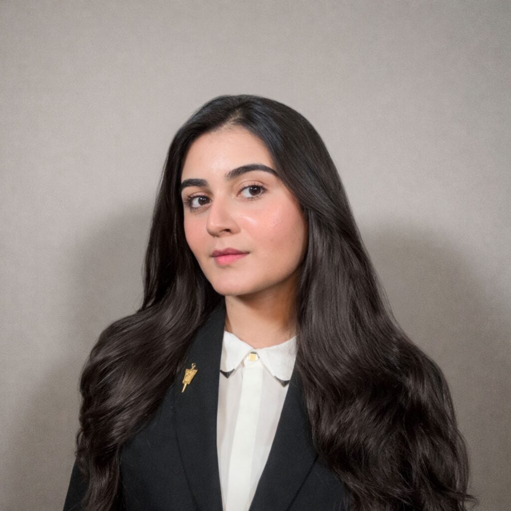 A woman with long, dark hair wearing a black suit jacket, white collared shirt, and a pin stands in front of a plain, light background, looking confidently at the camera.