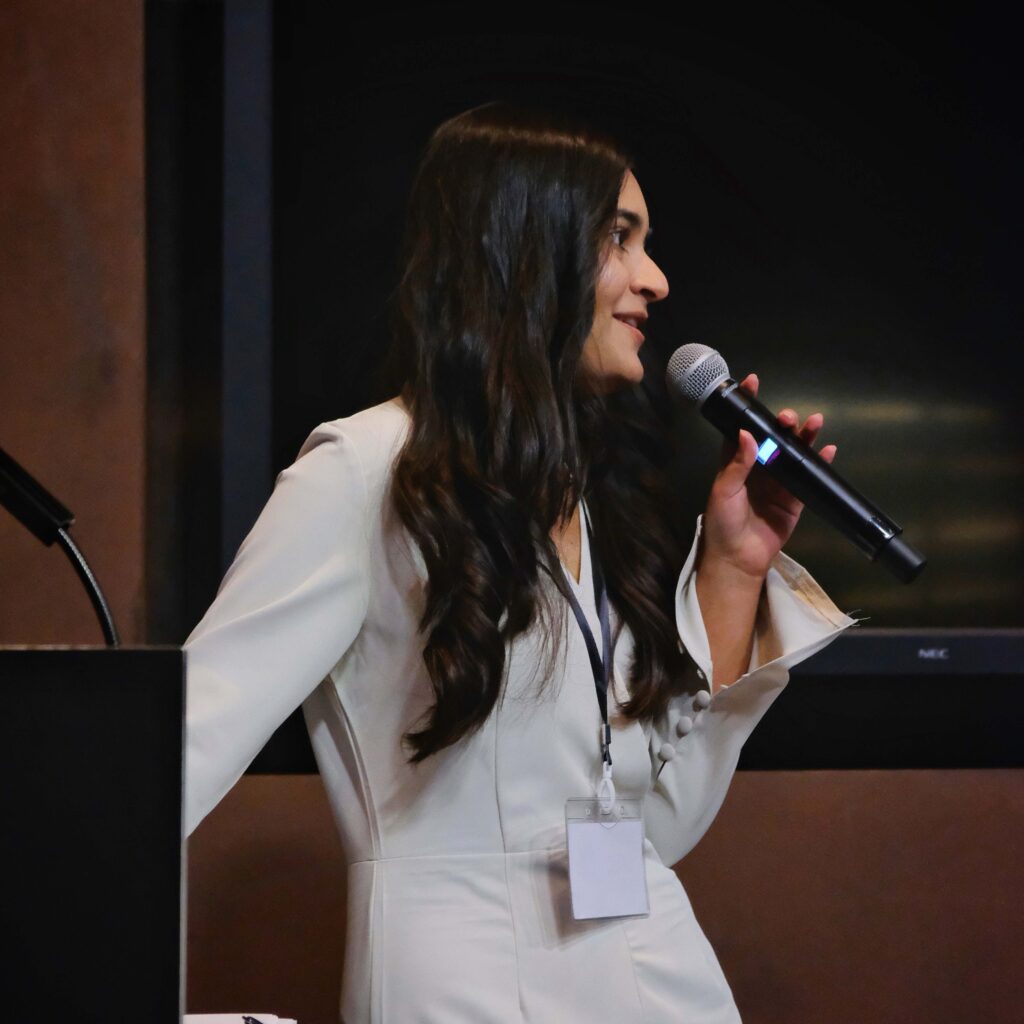 A woman with long dark hair, wearing a white outfit and name badge, speaks into a microphone while standing near a podium.