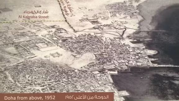 Aerial black-and-white photo of Doha, Qatar in 1952, featuring labeled Al Kahraba Street, a coastline, clustered buildings, and sandy terrain, with English and Arabic captions at the bottom.